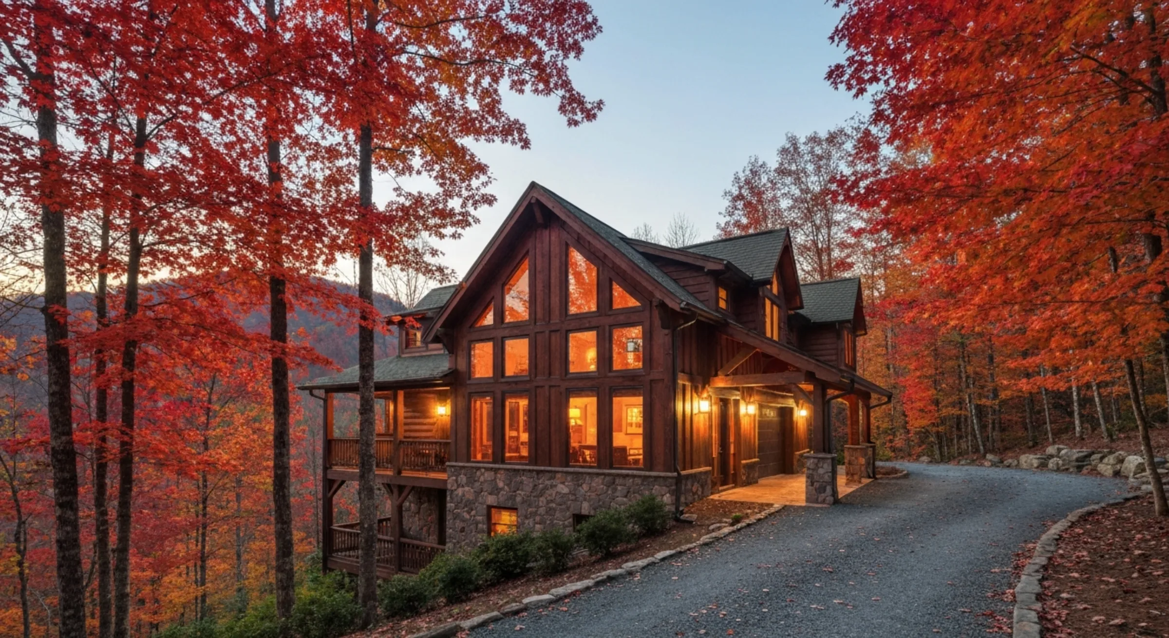 A wide editorial shot of a luxury Smoky Mountain short-term rental cabin in October peak fall foliage — wood-and-stone two-story timber-frame cabin with large picture windows glowing warm at dusk, surrounded by red and orange maple trees in vivid autumn color, gravel drive curving toward the entry. Real STR property listing photography.